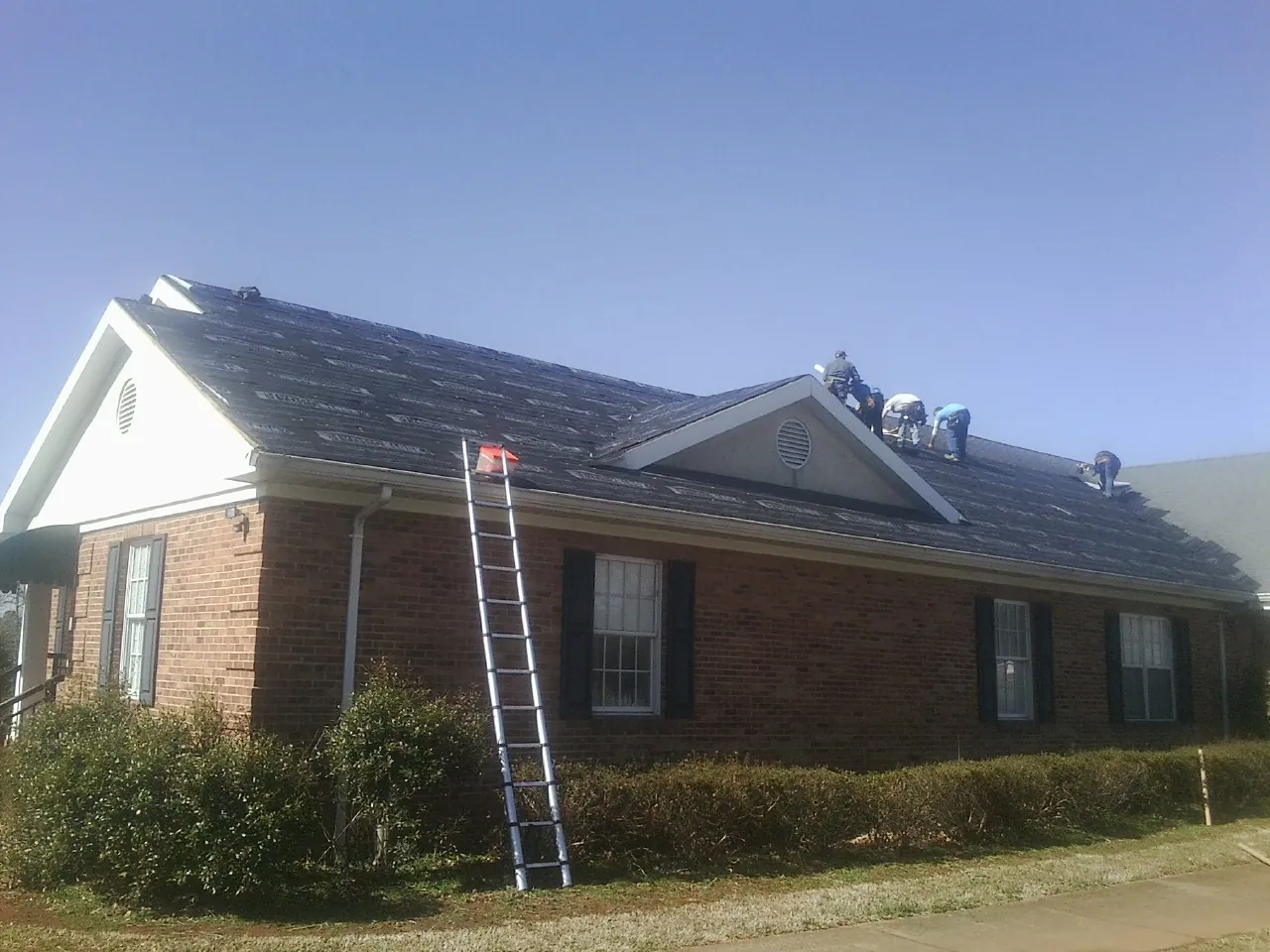 Crew tearing shingles off a brick ranch roof with a ladder set — Emergency Roof Repair work by our crew in Logan